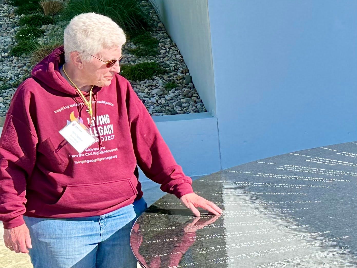 White woman with white wavy short hair and glasses, wearing a burgundy sweatshirt and blue jeans, touches a name engraved on black granite that has water on top of it