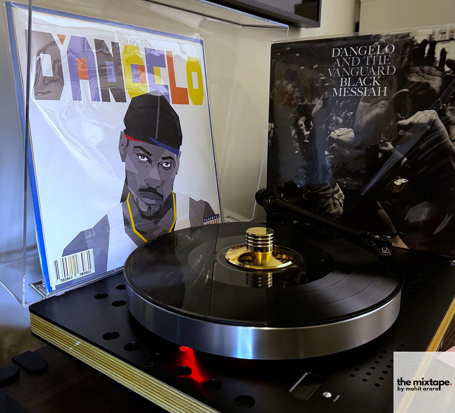 The image features a turntable in the foreground playing a vinyl record, with the album Black Messiah by D'Angelo and the Vanguard prominently displayed in the back right. The album cover showcases a grayscale photograph of a crowd in motion, evoking themes of activism. On the left, another album cover with stylized artwork of D’Angelo is visible but not the focal point of the composition. The setup highlights the appreciation for vinyl records and the music of D'Angelo.