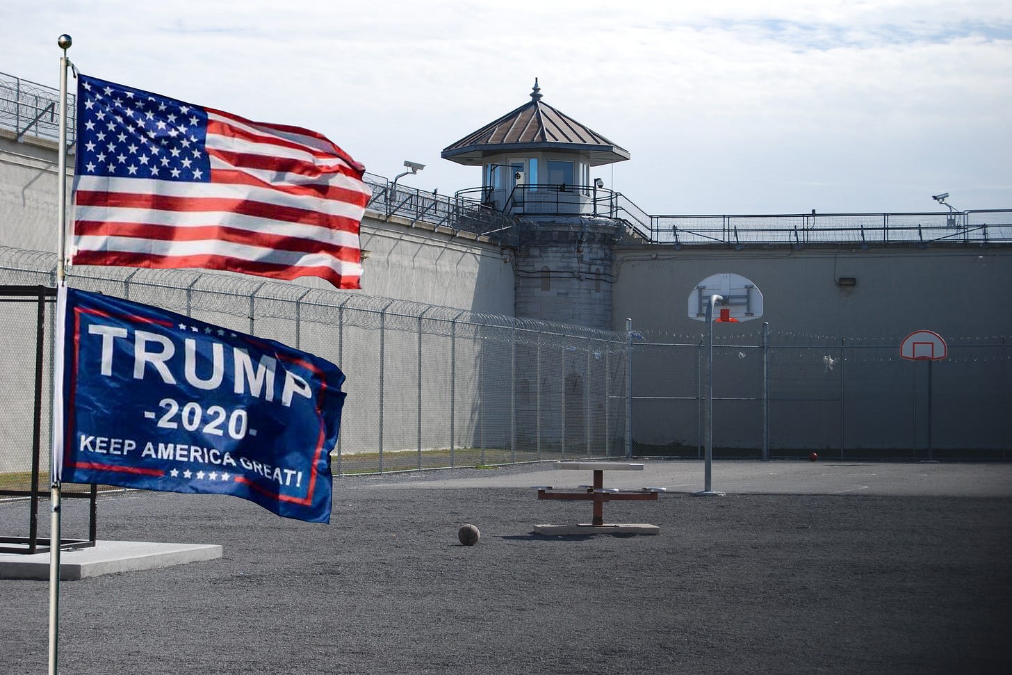 A photo of an empty prison recreation yard with a flag pole displaying the American flag and a Trump 2020 flag in the foreground. A photo of an empty prison recreation yard with a flag pole displaying the American flag and a Trump 2020 flag in the foreground.