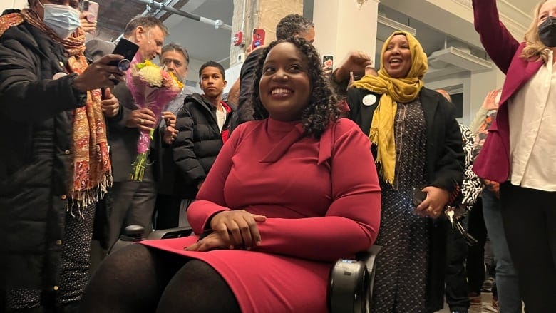 A Black woman, sitting in a wheelchair, smiles while people around her cheer and take photos. 
