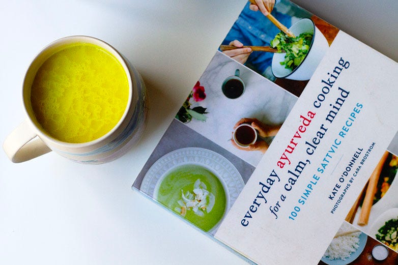 Turmeric milk in a ceramic cup with a copy of Everyday Ayurveda cookbook for a calm, clear mind : 100 simple sattvic recipes by Kate O'Donnell, against a white backdrop.