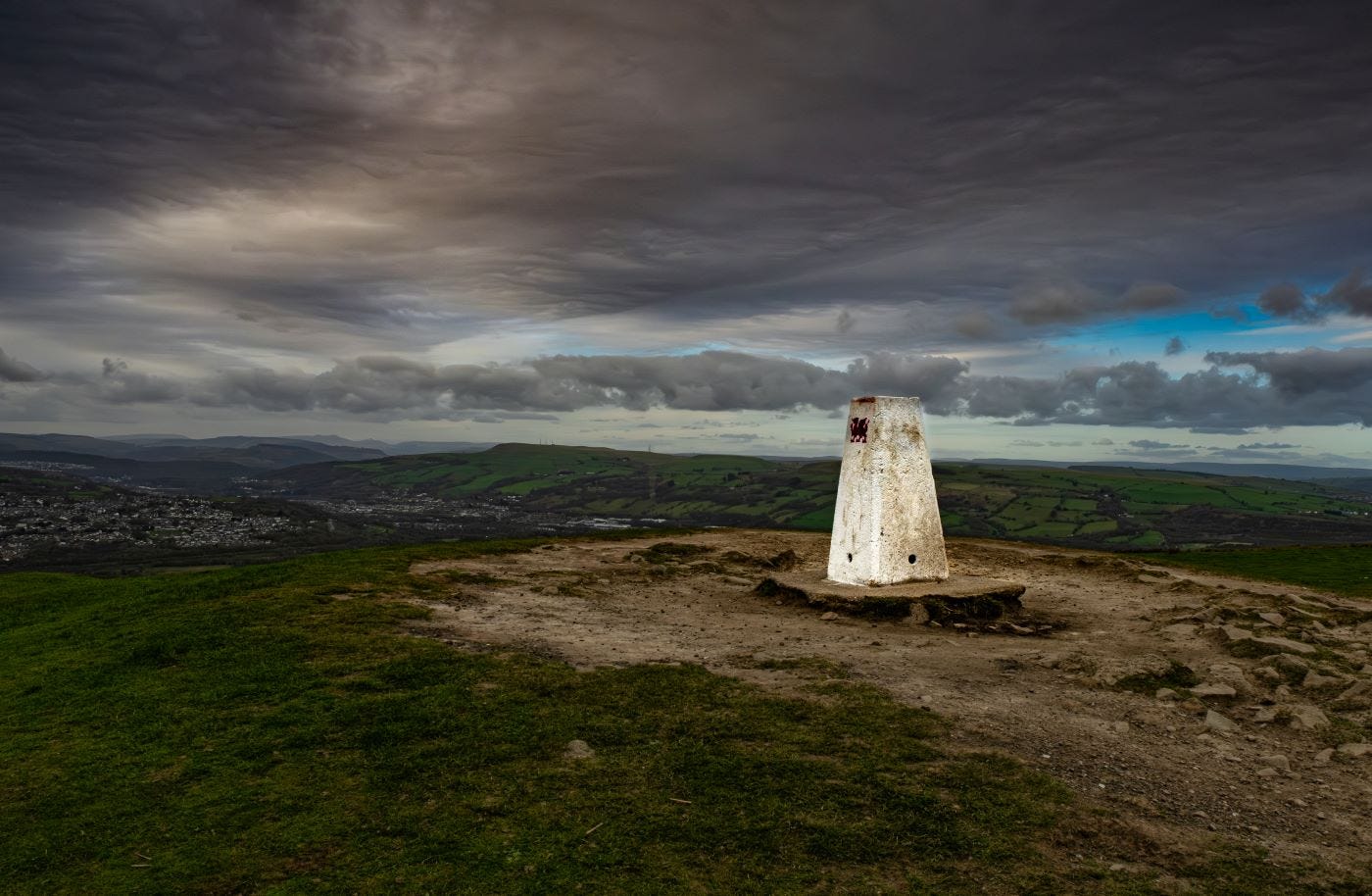 A trig point, which is a concrete block about four feet high and painted white, stands on top of a hill. The green grass of the hill is worn away around the trig point, exposing bare earth. A red dragon is stencilled on one side of the trig point. The sky is cloudy, dark and brooding.