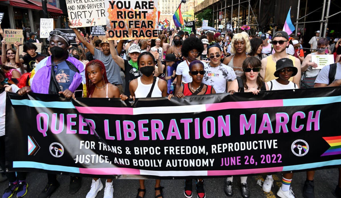 A crowd of people at a protest hold signs as they march. The front line of eight people, some wearing masks, are holding a banner that reads 'Queer Liberation March for trans & BIPOC freedome, reproductive justice, and bodily autonomy. June 26, 2022'. A person directly behind this row holds a sign that reads 'You are right to fear our rage'.