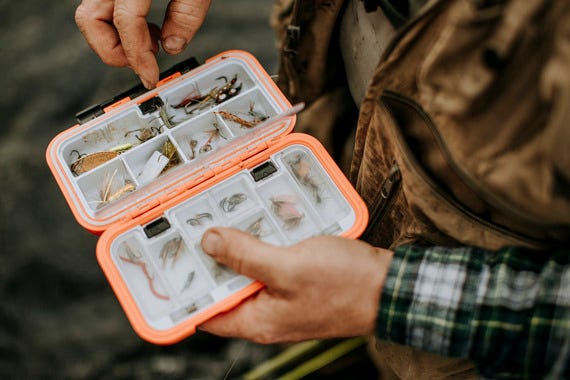 fly angler looking through fly box wearing a tan fly fishing vest and flannel shirt