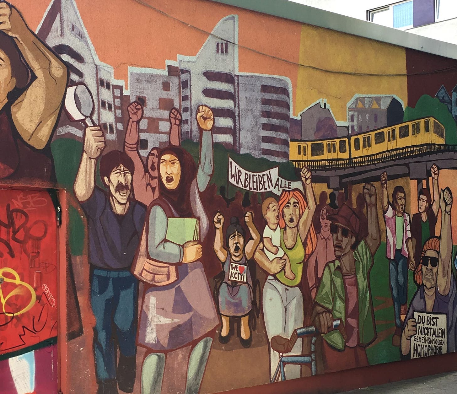 A photo of a mural painted on a wall in the Kotti neighborhood of Berlin. The mural shows buildings, a train and people protesting with sights that say Wir Bleiben Alle, which means We All Stay in English.