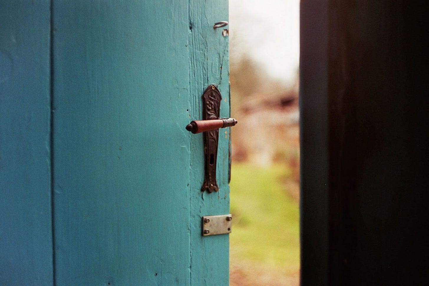 A teal, plank wood door with an ornate, antique handle open just a bit to see an out of focus green path outside. A teal, plank wood door with an ornate, antique handle open just a bit to see an out of focus green path outside.