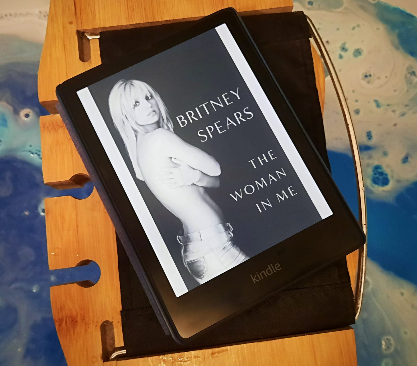 Paula’s Kindle is on a wooden tray placed over a bath. The Kindle is open on the title page of The Woman in Me by Britney Spears. The bathwater is blue as the result of a bath bomb.
