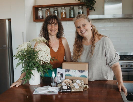 Image of Cat Green and Heidi Merika standing behind DIY Immunity Remedy Kit with Kit items displayed in front of it - herbs, bottles, recipe cards etc. herbal medicine education made simple and fun for family herbalists and home herbalists. Everyday Empowered