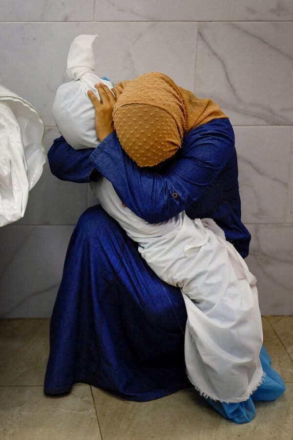 A woman in a brown head scarf and blue robe embraces the body of a child wrapped in a sheet.