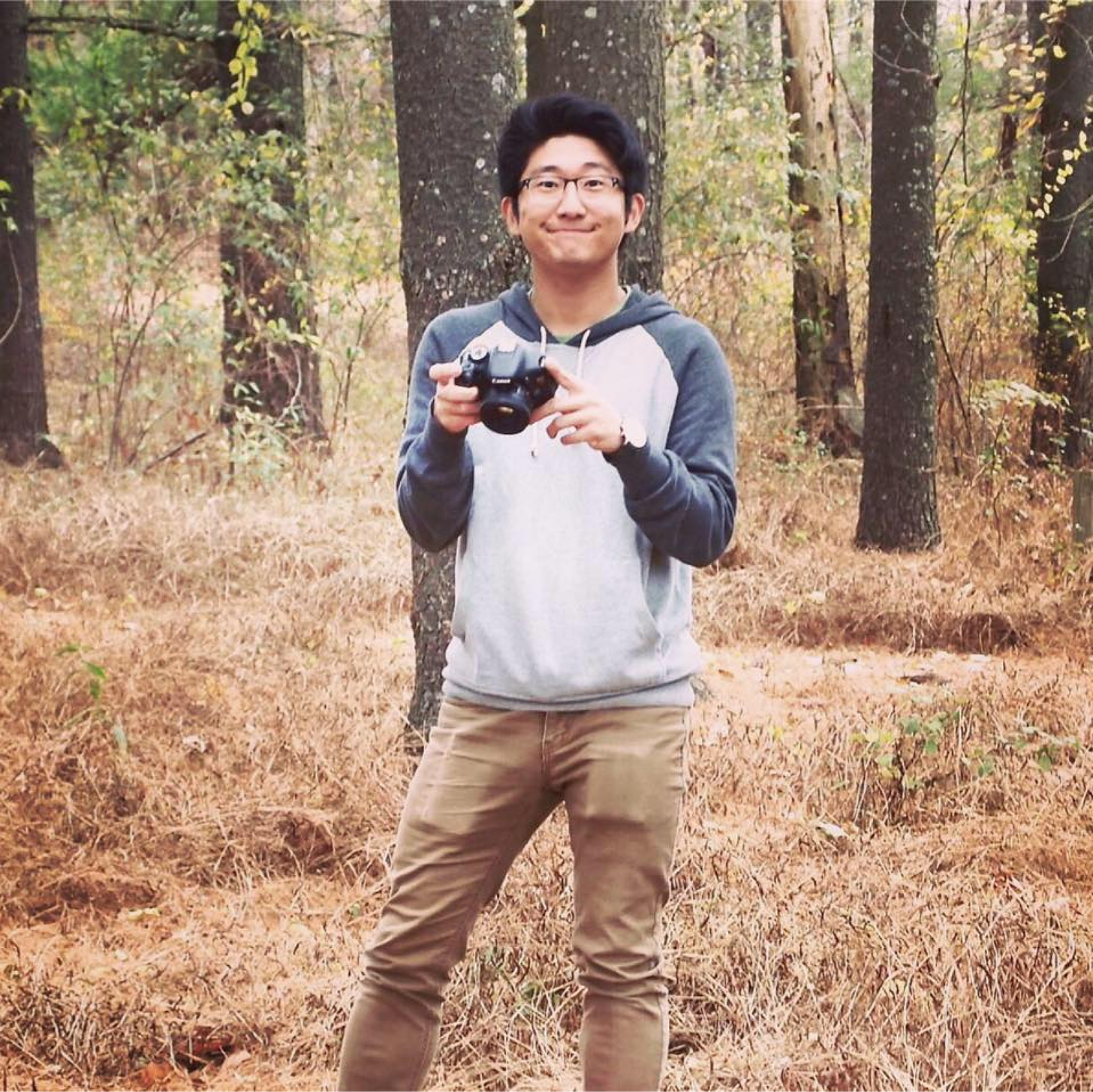 Sam smiling while holding his DSLR camera standing in the middle of a field with trees in the background.