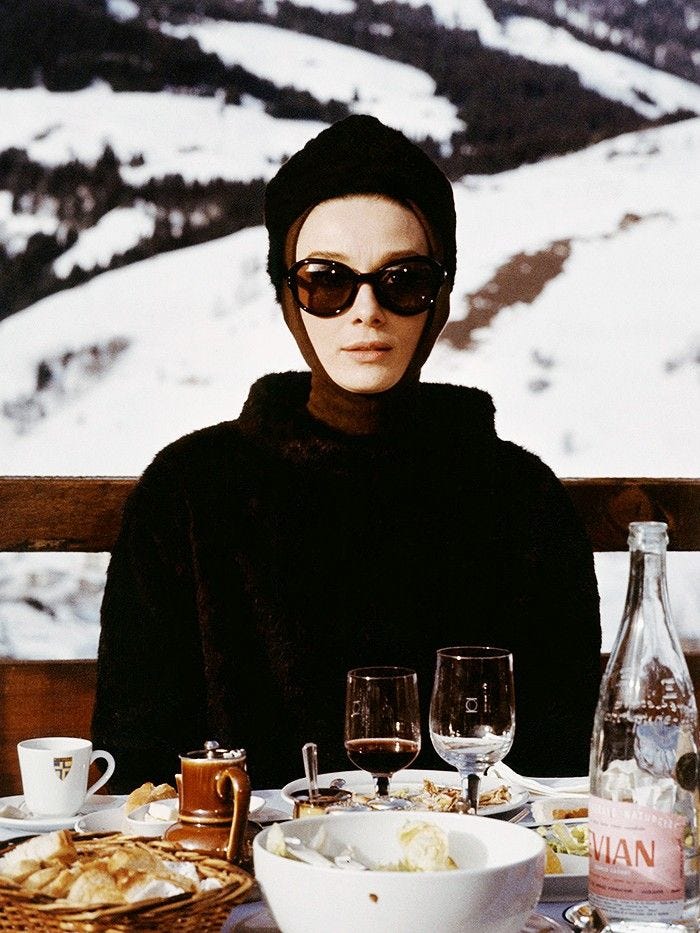 Audrey Hepburn wearing an all-brown Givenchy brand ski outfit, including a brown fur coat and brown sunglasses, seated at a table at a ski resort with breakfast on the table in front of her, snow-covered slopes are seen in the background