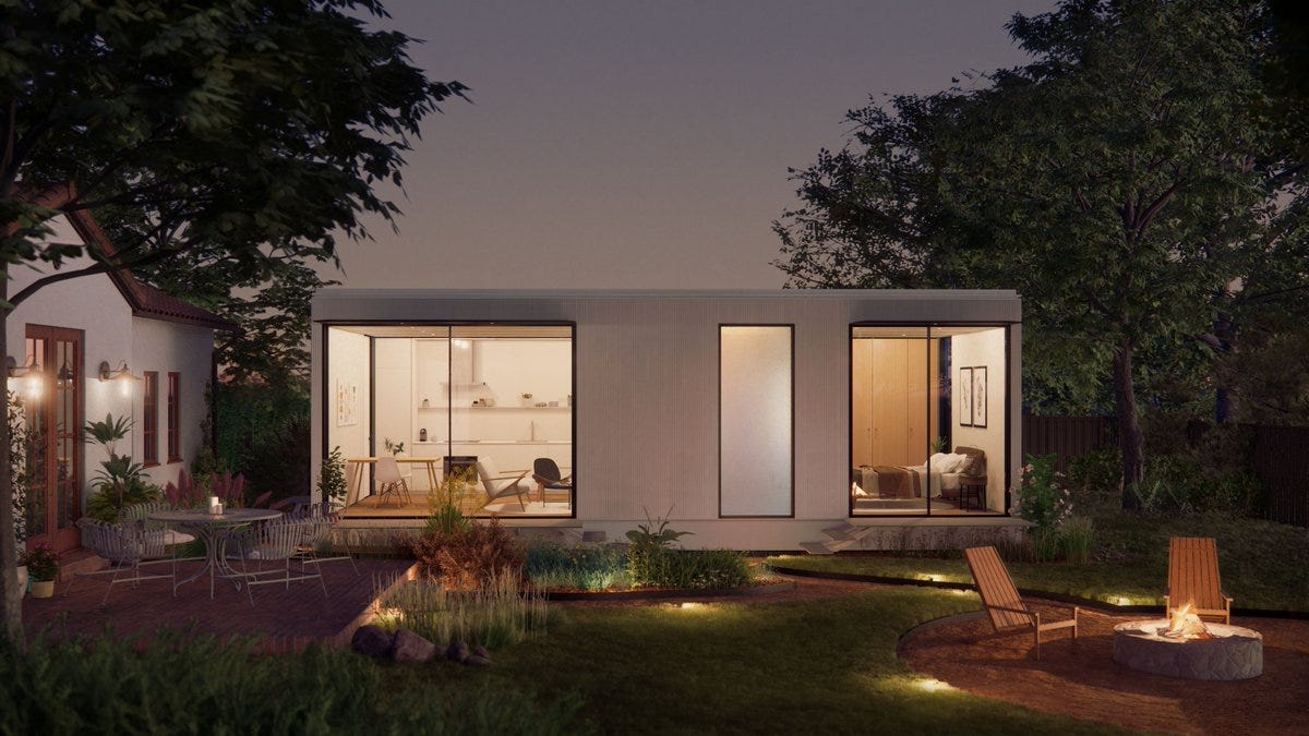 This modern, rectangular single-story prefab house boasts large windows and is nestled in a lush garden. A patio set graces the left, while a lit fire pit with chairs offers warmth on the right under the serene evening sky.