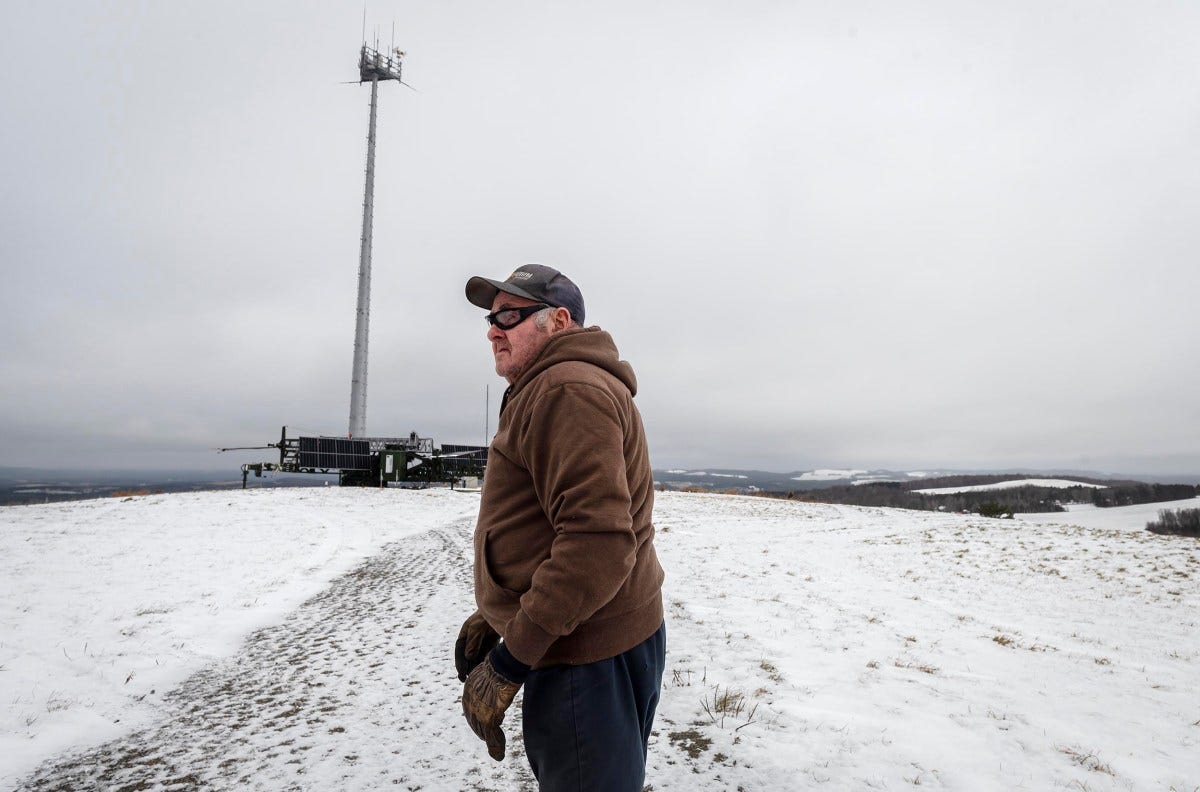 A man in a brown jacket and cap stands on a snowy hill near a tall communication tower under a cloudy sky.