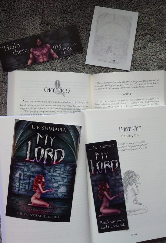 A photo showcasing the internal layout of the book, with the artwork & header art, a bookplate showing a sketch of the front cover, bookmarks with the cover art, 1 with Meya stating "break the cycle & transcend", the other with Lord Deminas stating "hello there, my pet." Image description of the front cover: The background is that of a dungeon in grey & blueish hues. In the upper part of the cover it reads L. B. Shimaira, & below that My Lord. The title lettering appears in a silver metallic font with red & purple blood dripping from the bottom. In the lower half of the cover a naked white woman with wavy red hair is seated on her knees, facing right. She has her hands clasped together in front of her & blood is spilling from them into a chalice by her knees. Her eyes are closed, & her chin is slightly turned up. The floor around her is covered in red blood that slowly changes to purple as it runs off-screen towards the viewer. To the left of her, on the wall, are shackles.