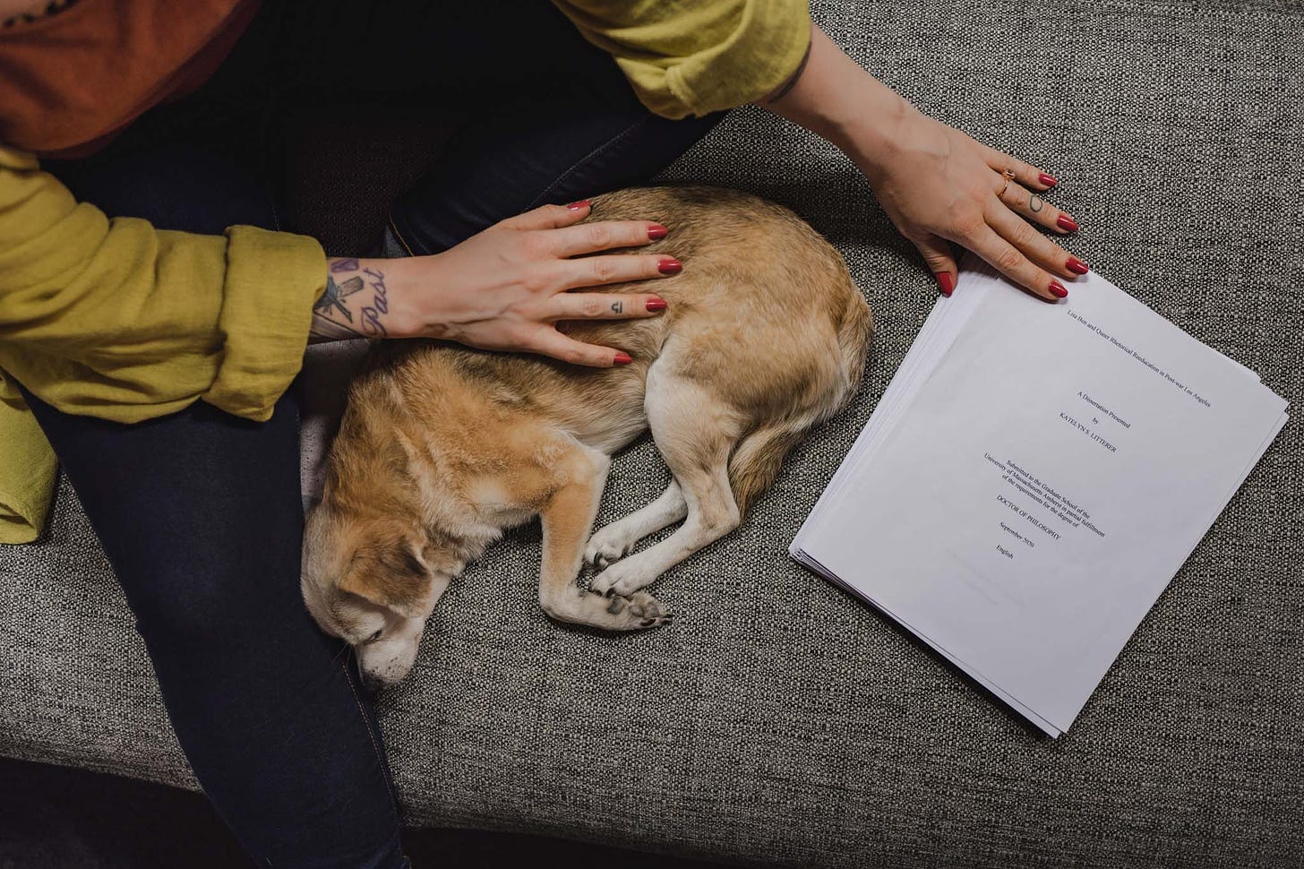 Image Description: Kate is sitting on a couch snuggling with her sleeping dog, Lucy, with her hand on her printed dissertation.