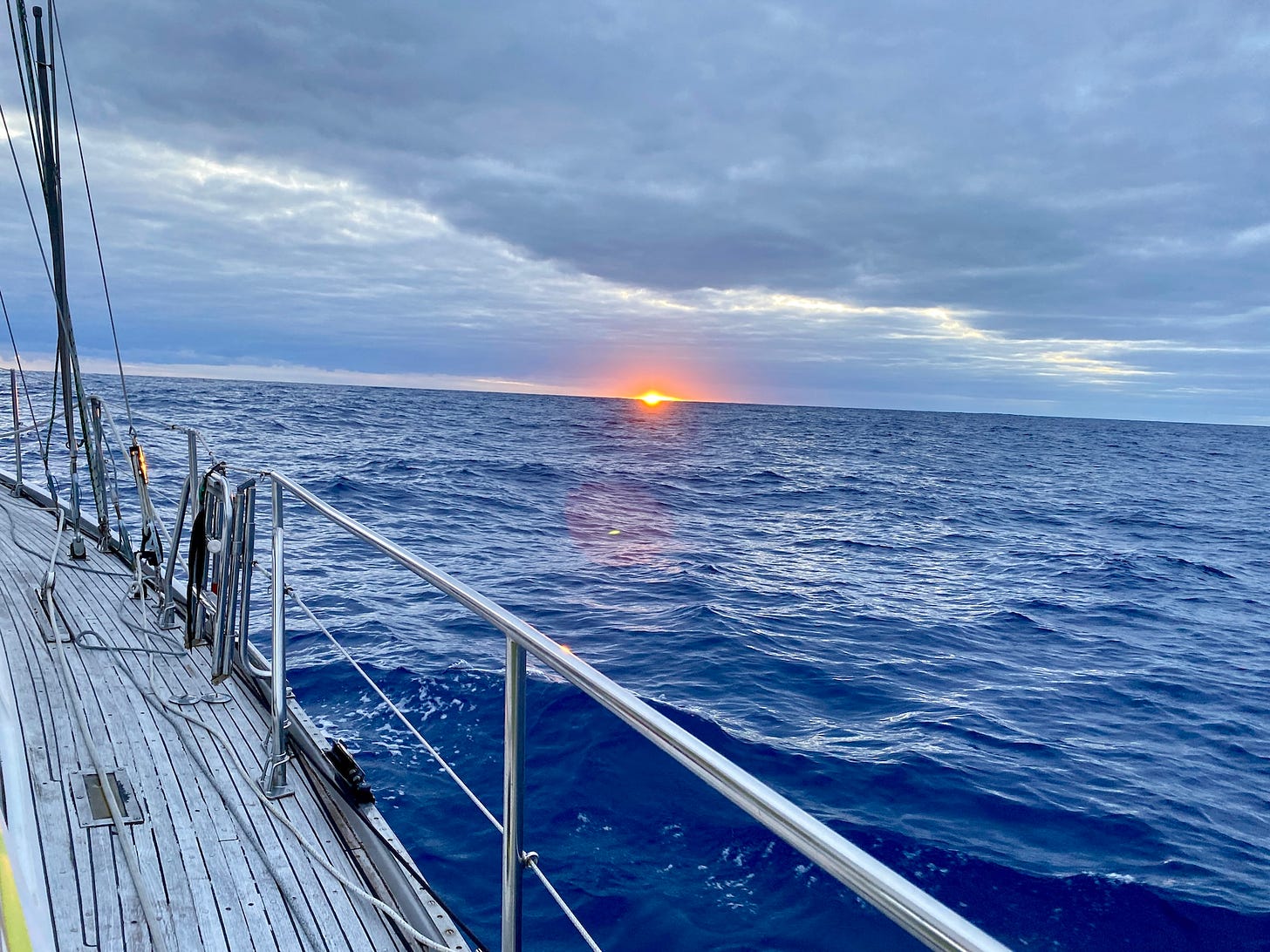 A view of the sunrise on the ocean. The deck and rails of a sailboat are in the foreground, the sun is brilliant orange at the horizon.
