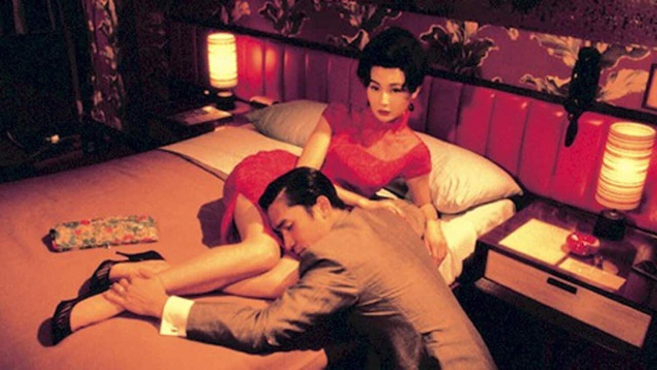 A woman in a red dress reclines on a bed while a man, kneeling on the floor, rests his head against her and caresses her ankle.