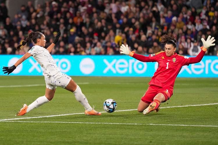 In a crowded soccer stadium, a Black woman in a white shorts-and-tee uniform is in action kicking the blue-and-white swirly soccer ball, while a goalie, a woman with long dark brown hair in a ponytail, wearing a red uniform and big white gloves, is sliding towards the ball to block it.