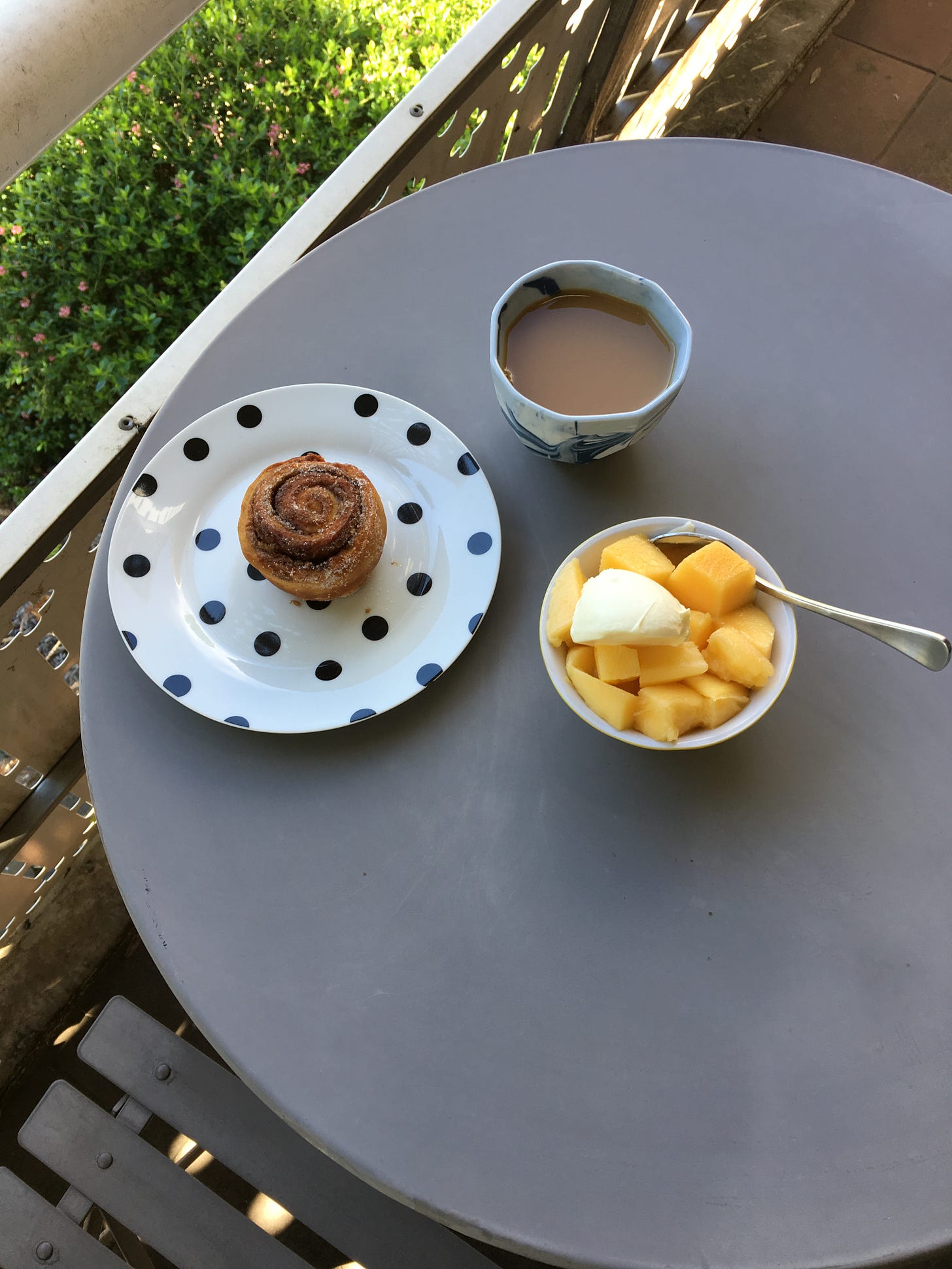 Cinnamon scroll on a spotty plate alongside milky tea and a bowl of mango and yoghurt
