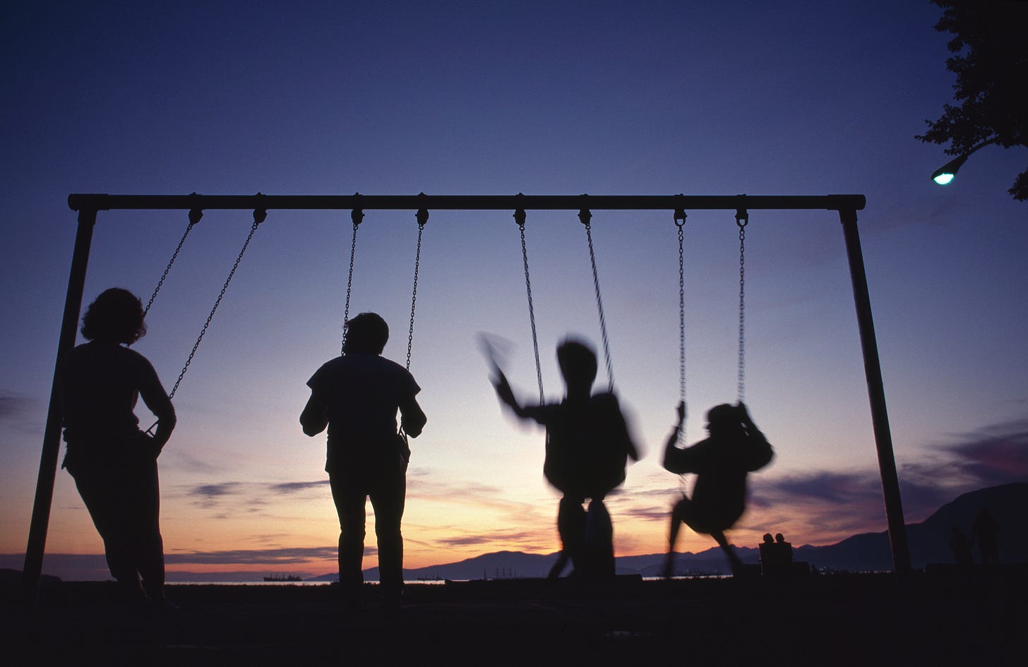 Four people enjoy playground swings at dusk.