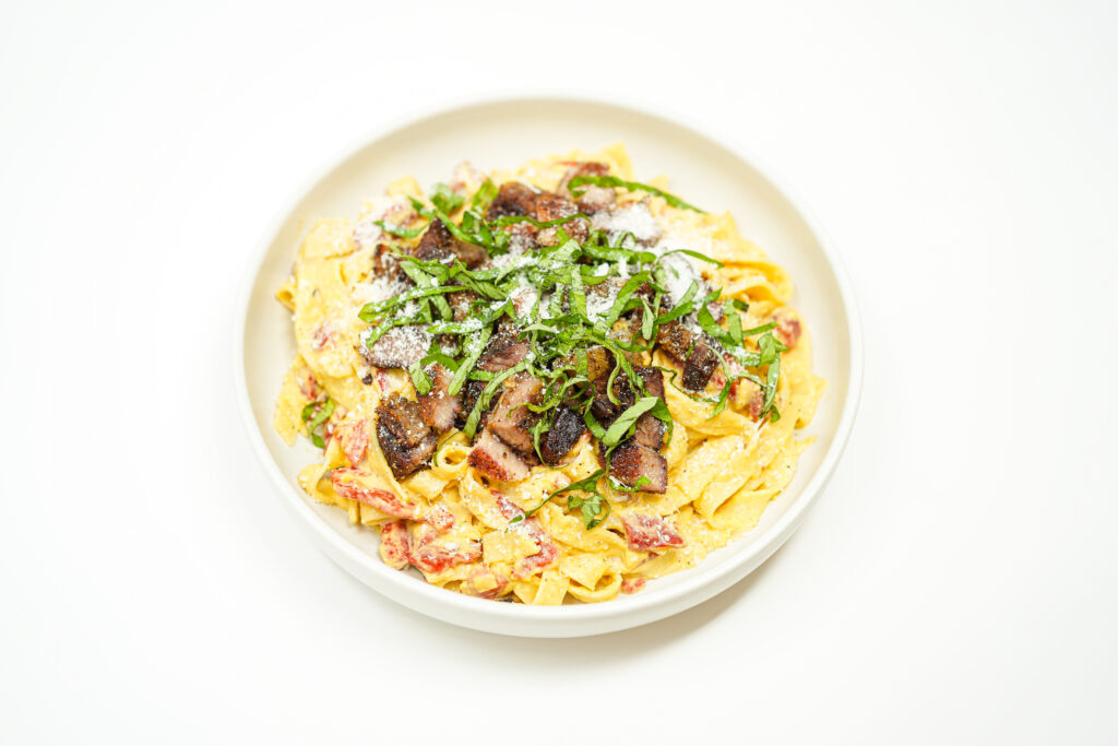 A plate of crispy pork belly pieces served over creamy corn-coated tagliatelle, with sautéed red bell peppers, fresh herbs, grated Parmesan, and lemon zest.