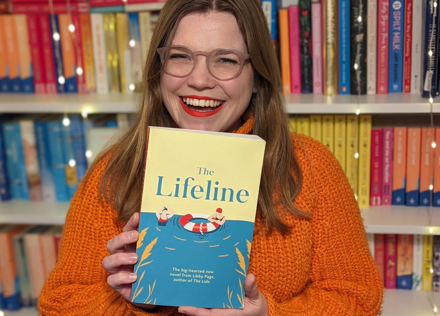 Author Libby Page is wearing an orange jumper and smiling while holding a copy of her book, The Lifeline. There are bookshelves in the background.