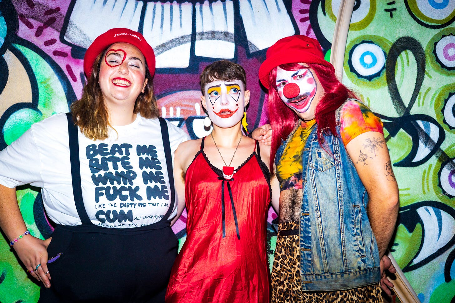 L-R: Caty, Cass, and Babz pose in clown outfits and makeup against a colorful graffiti wall