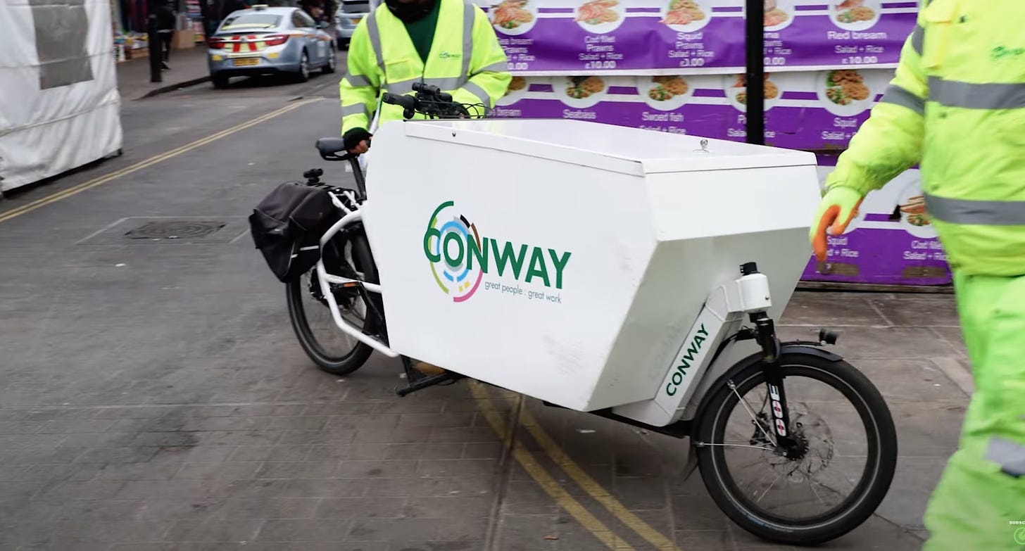 A modern cargobike branded with Conway logo. A cycling delivery rider walks the bike into a building site to deliver an item.