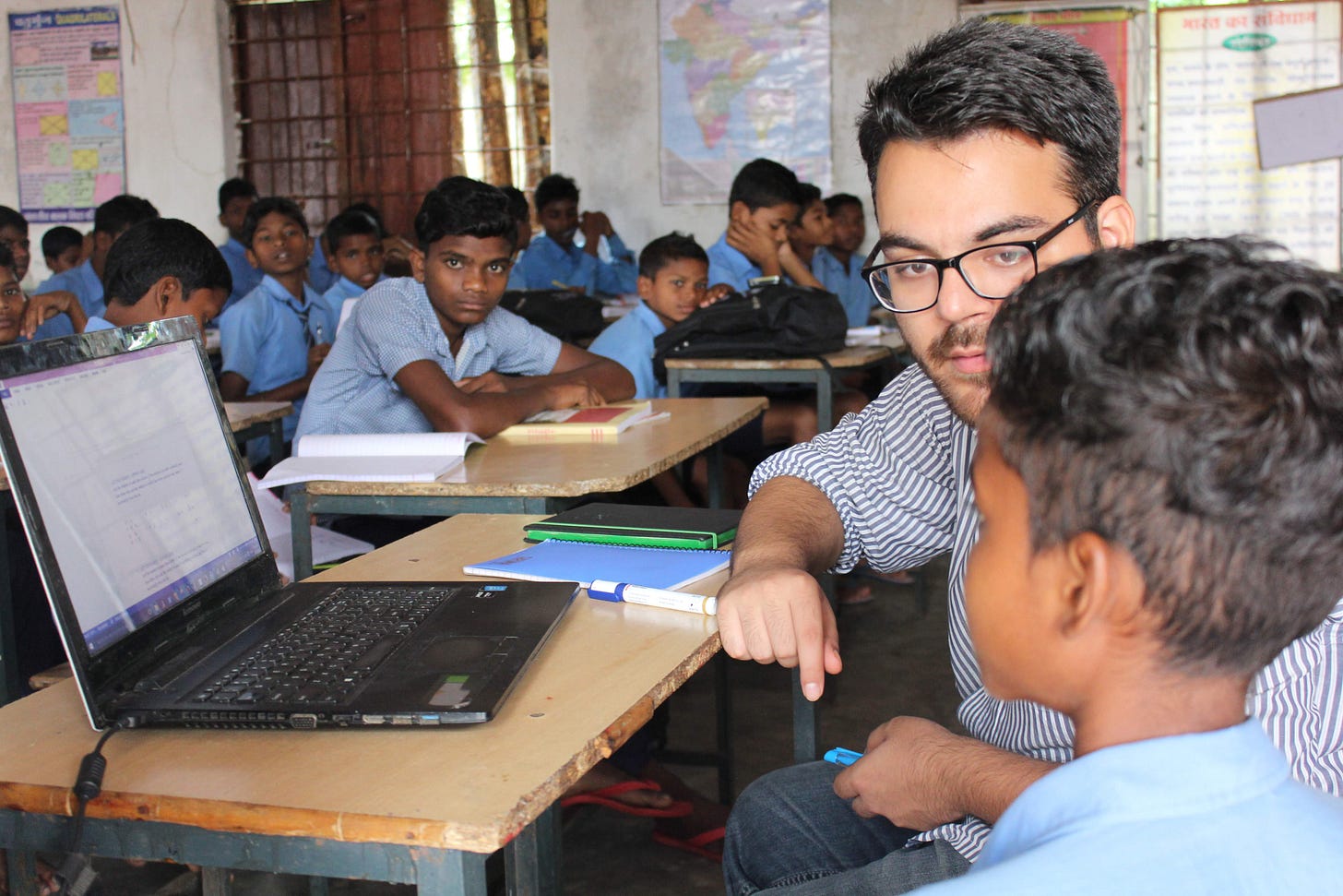 A man teaches a child something on a laptop, possibly- how to use a laptop. In the background is a classroom full of children A man teaches a child something on a laptop, possibly- how to use a laptop. In the background is a classroom full of children