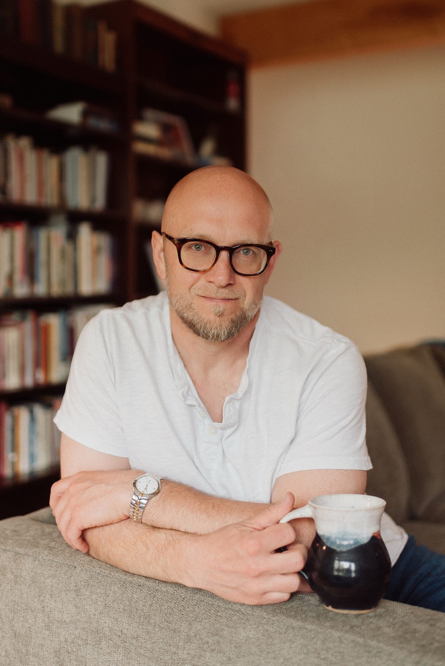 A bald man with a beard and glasses is leaning on the back of a couch, holding a ceramic mug. He is wearing a white short-sleeved henley shirt and a silver watch on his left wrist. He is smiling slightly and looking directly at the camera. In the background, there is a bookshelf filled with books, suggesting a cozy and intellectual setting.