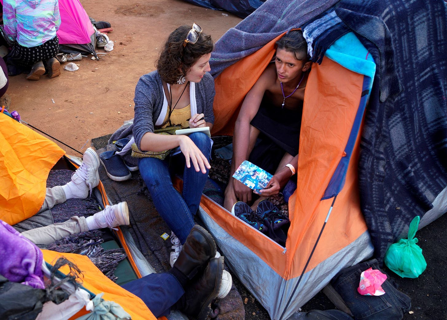 A woman journalist sits outside a tent and speaks with a young man who sits inside the tent