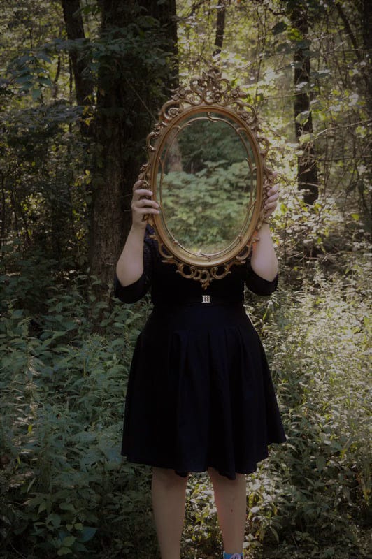 Person in a dress holding an antique mirror blocking their face and reflecting a forest.