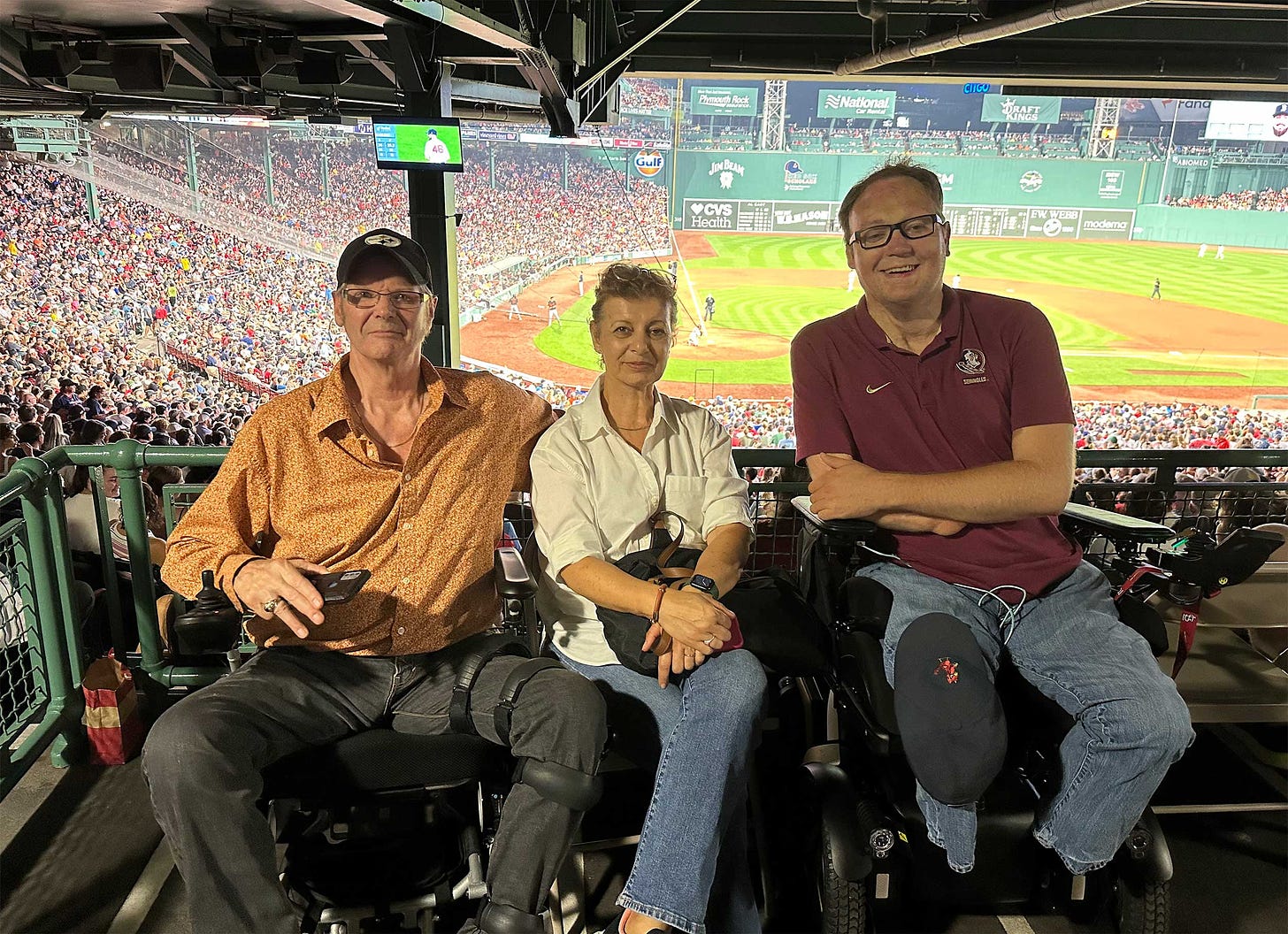 John seated in his wheelchair next to two readers inside baseball park.