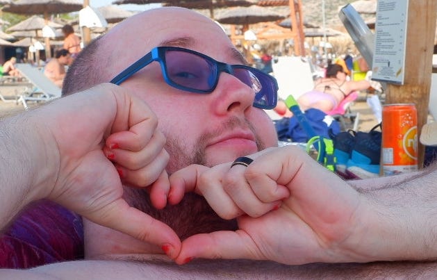 A close-up photo of an agender person (me), on the beach, wearing blue glasses, and doing heart hands at the camera