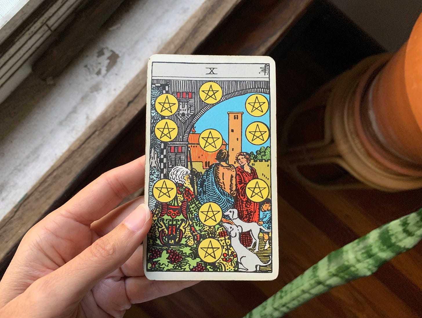 A hand is holding a tarot card, ten of pentacles by Pamela Colman Smith for the Rider-Waite-Smith deck. In the image, an old person with white hair is sitting down with two dogs near him, as two adults are speaking in the background with a child nearby as well. There are ten pentacles dispersed throughout the card in the shape of the kabbalistic tree of life. Behind the card is an open window sill, a wood floor and a terra cotta pot with a house plant in it that is sneaking into the photo just slightly.