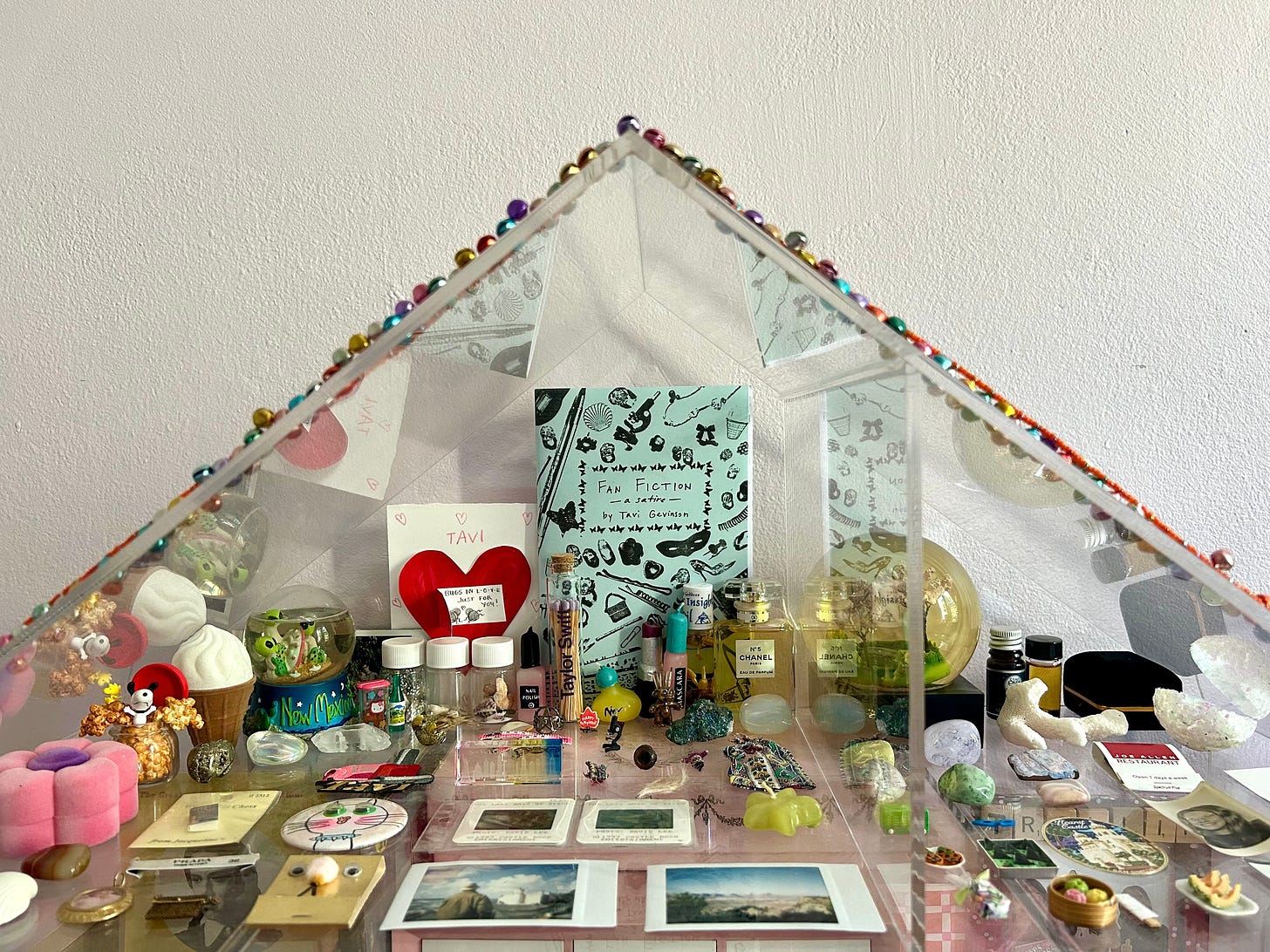 Photograph of a clear acrylic shelf that is decorated with various neatly arranged objects, like a shrine. Objects include polaroid photographs, lipstick, matchboxes, a snowglobe, crystals, and other knick-knacks. A copy of the zine “Fan Fiction” is prominently displayed in the center-back of the shelf. 