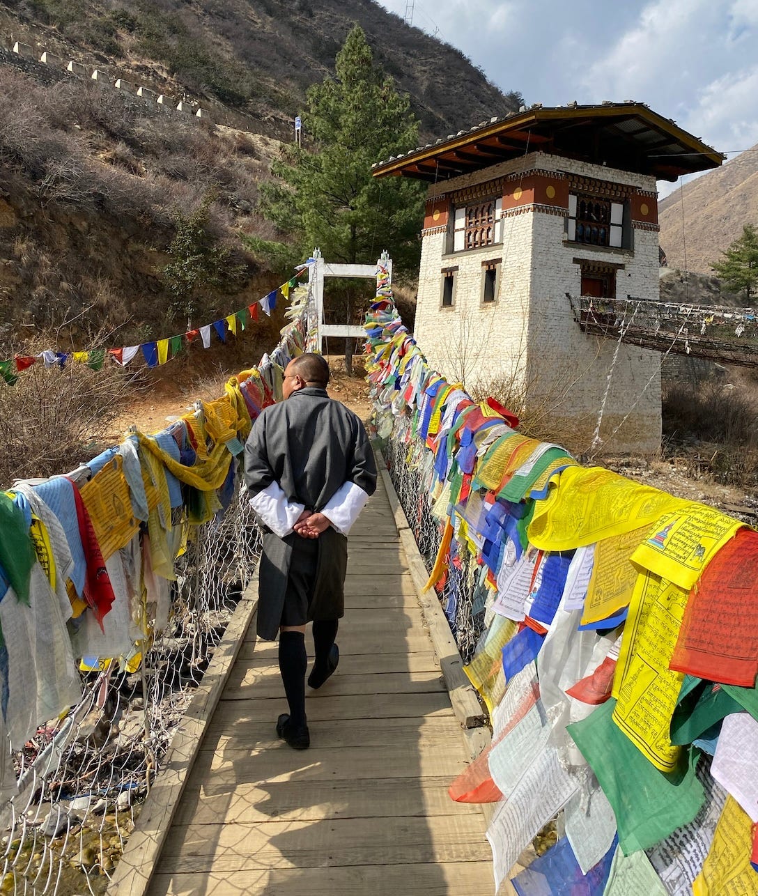 My guide Kahar, dressed in a grey Gho, a traditional Bhutanese knee-length cloth tied at the waist, walking across a bridge covered in colorful prayer flags