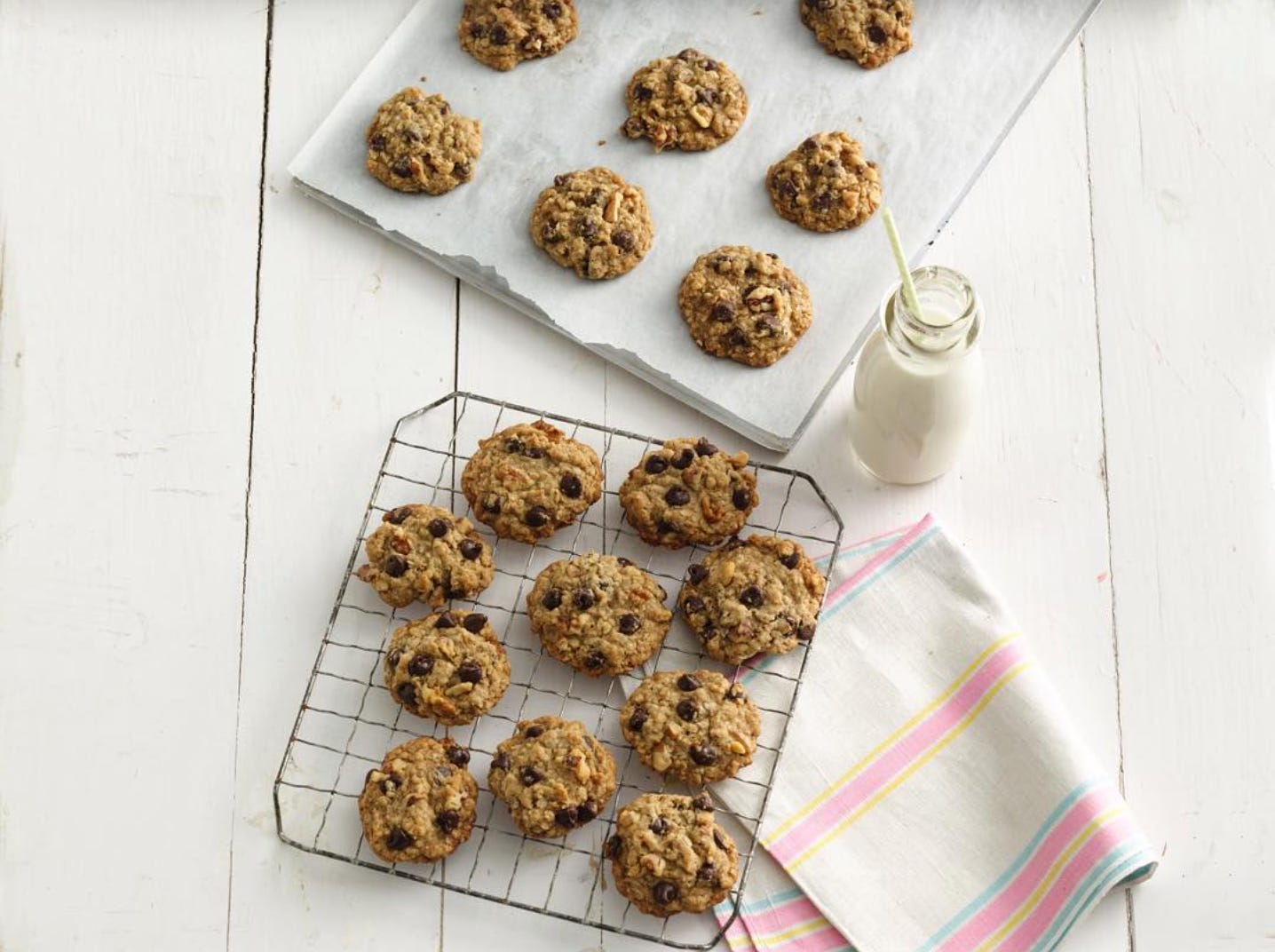 Image shows freshly baked cookies cooling on a baking sheet and a cooling rack plus a glass bottle of milk with a straw in it.
