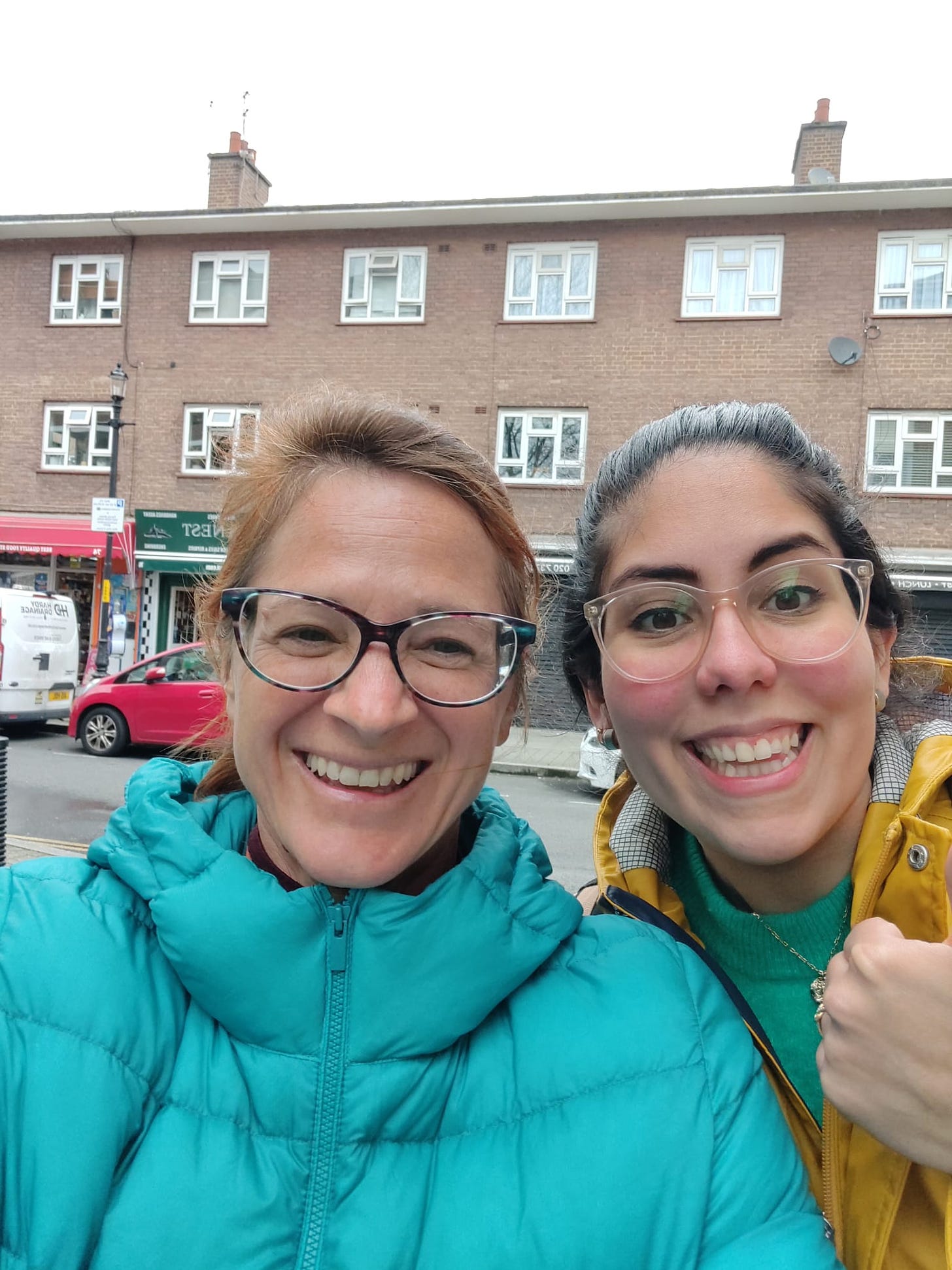 A portrait of Yanna and Alejandra smiling both with a ponytail and glasses in a street in London with shops in the background