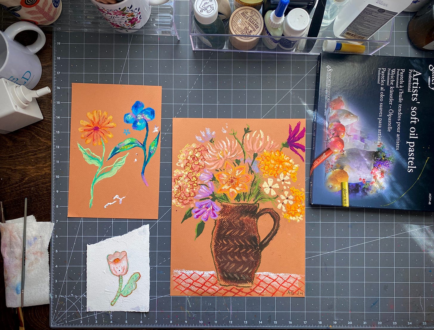 A photo of the artist's studio desk with art supplies, focusing on three different oil pastel flower still life drawings.