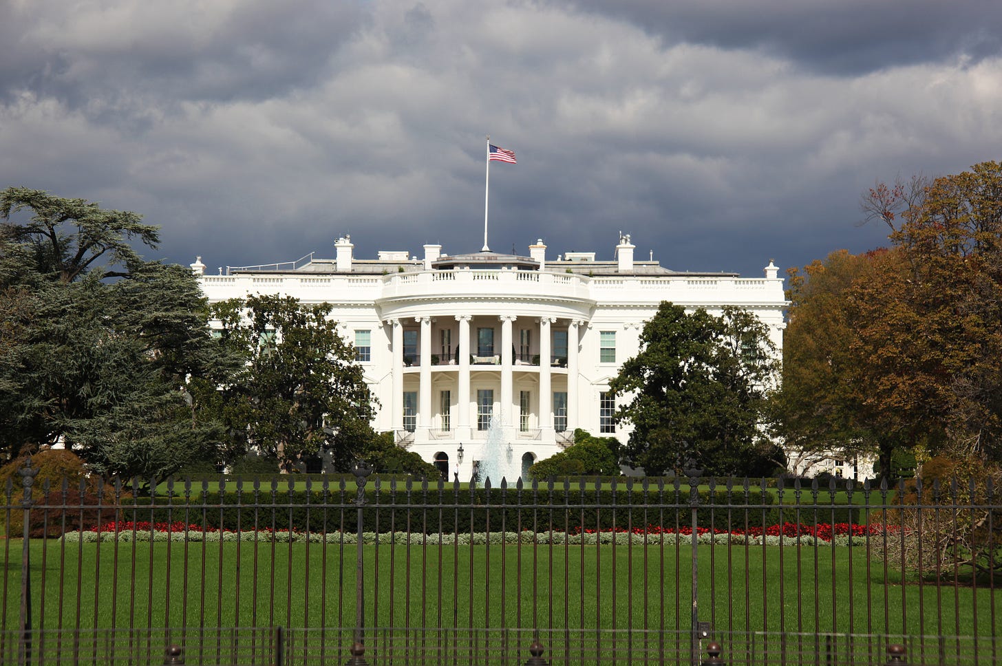 Close up photo of the White House with green grass and trees around it and low, dark clouds hanging over it. The American flag is flying over top of it. Close up photo of the White House with green grass and trees around it and low, dark clouds hanging over it. The American flag is flying over top of it.