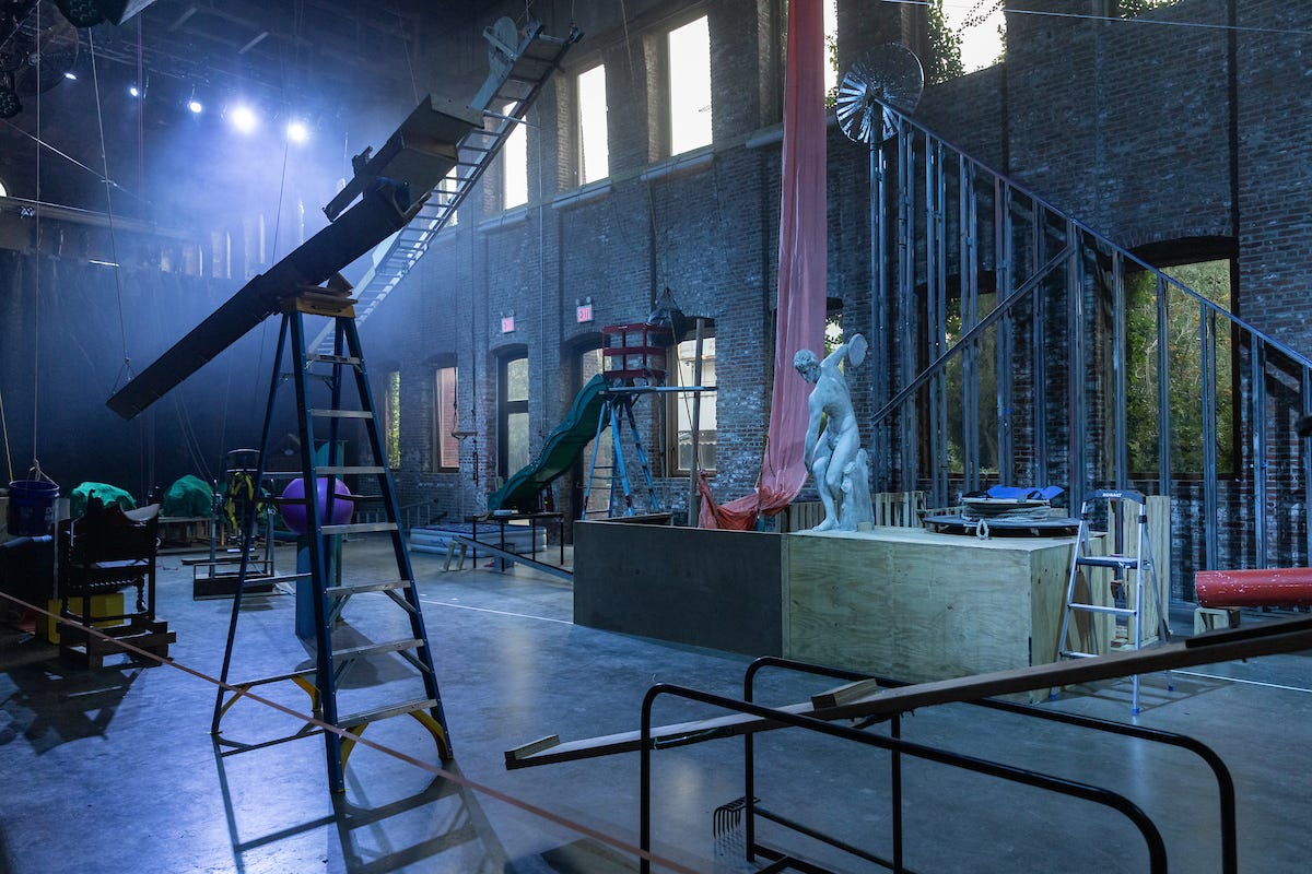 A brick-walled warehouse space filled with objects and contraptions like a see-saw, ladders, ropes, and a classical Greek statue.