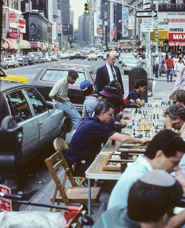 This may contain: a group of people playing chess in the middle of a busy city street with tall buildings