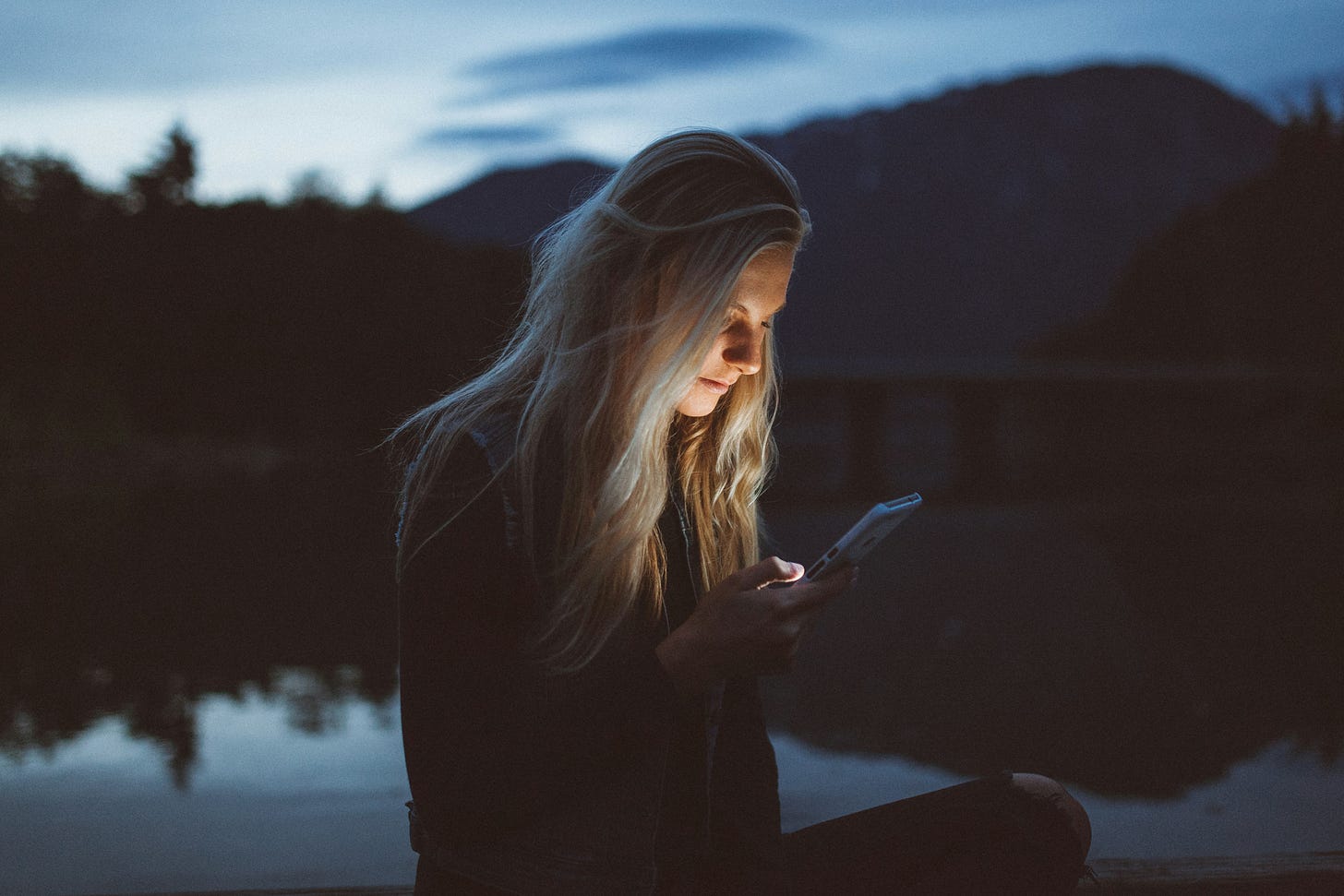 blonde woman in profile with face lighted looking at cell phone in almost total darkness