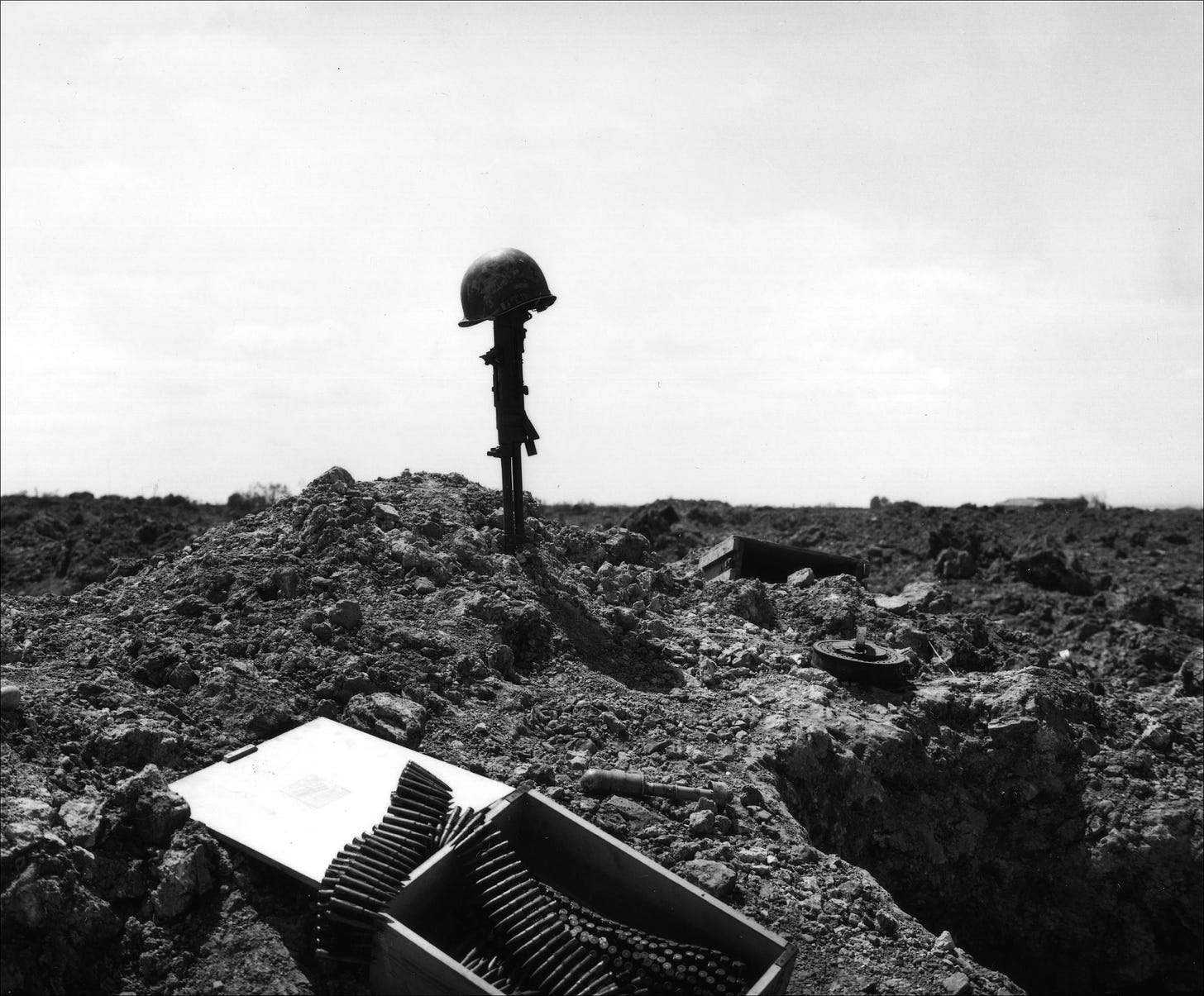 Black and white photo of a machine gun stuck into the ground, a GI helmet placed atop it, against a stark white cloudy sky. In the foreground are an open box of machine gun ammunition belts, an unexploded German grenade, and other debris of battle on the churned-up earth.