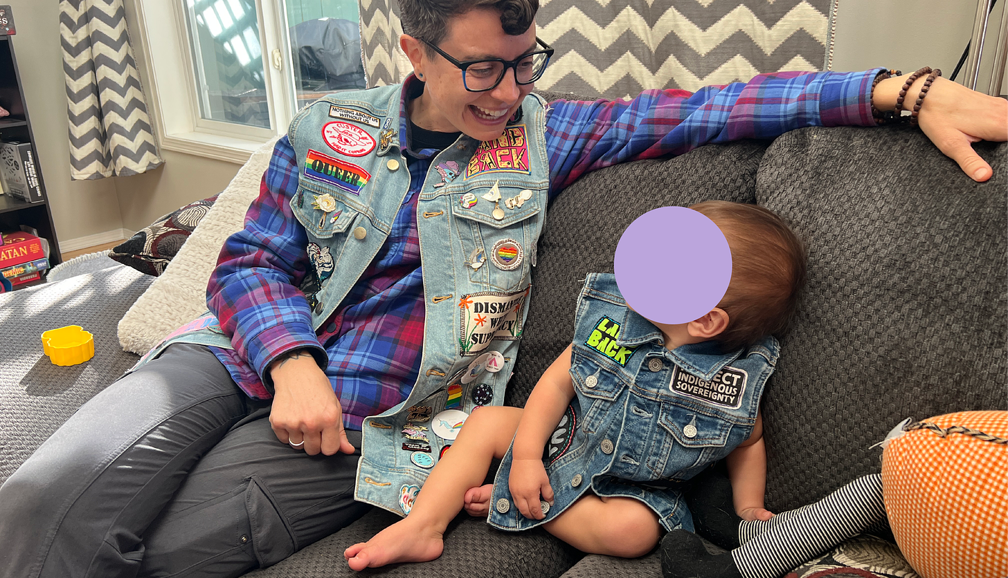 Photo of the Artist in their own Punk Rakusu worn over a plaid red and blue flannel shirt. They are sitting on a grey couch next to a one-year-old, also in a denim vest with patches on it. 