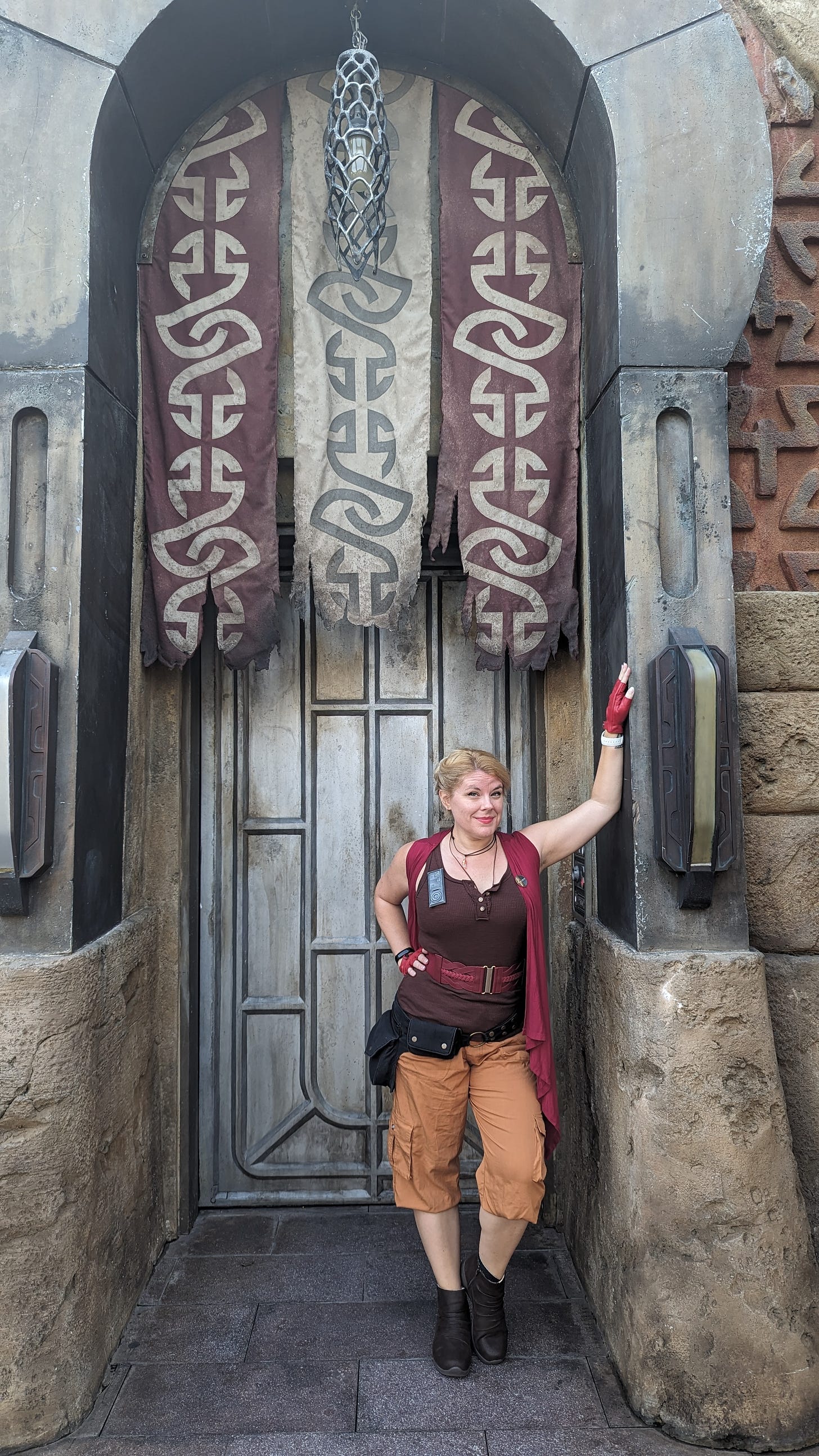 Cass, dressed in brown-orange cargo pants, a brown tank top, a red sleeveless wrap, and red gloves, standing in front of a decorative archway in Black Spire Outpost