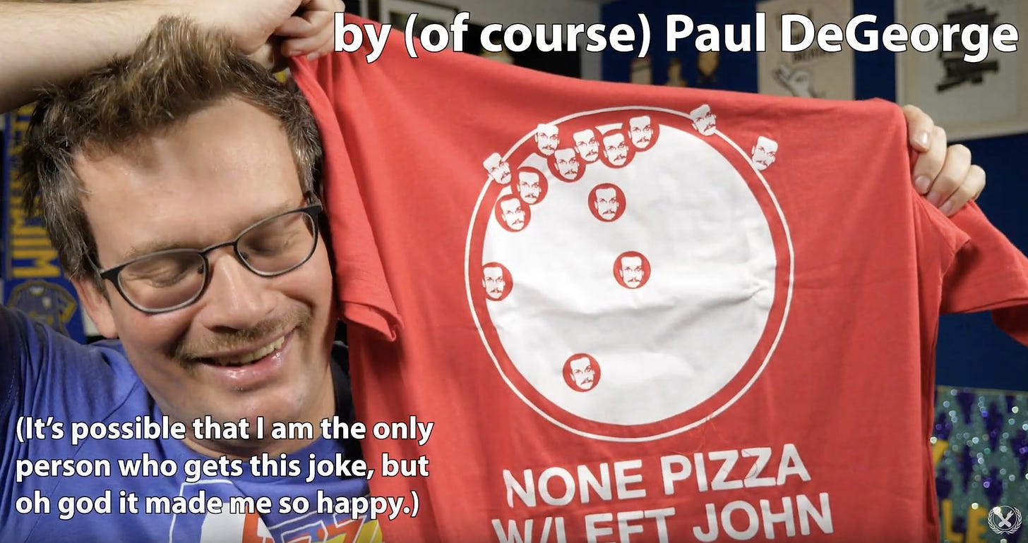 Screenshot from a YouTube video in which John Green holds up a reddish shirt with a picture of his face scattered mostly on one side of a circle, under which is printed "None pizza with left John."