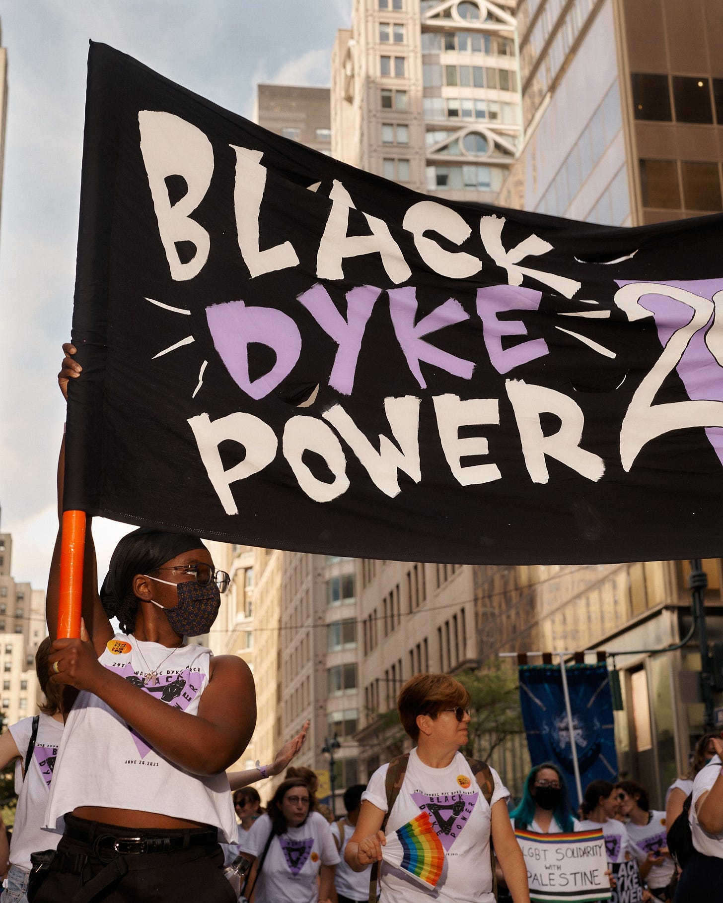 A Black person with a mask on holds up half a banner that says BLACK DYKE POWER A Black person with a mask on holds up half a banner that says BLACK DYKE POWER