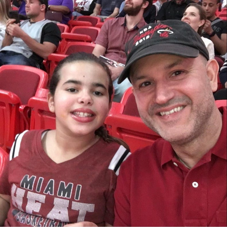 A young girl in a Miami Heat shirt sitting in the stands of a sports arena, next to a smiling man in a sports cap